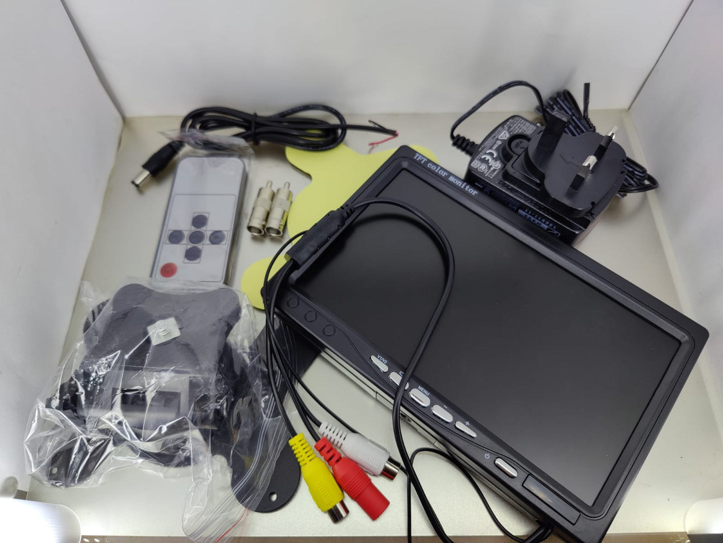 An unboxed 7-inch CCTV monitor is laid out with its accessories, including the mounting bracket, remote control, cables, and power adapter, illustrating the complete package for setting up a security surveillance display.