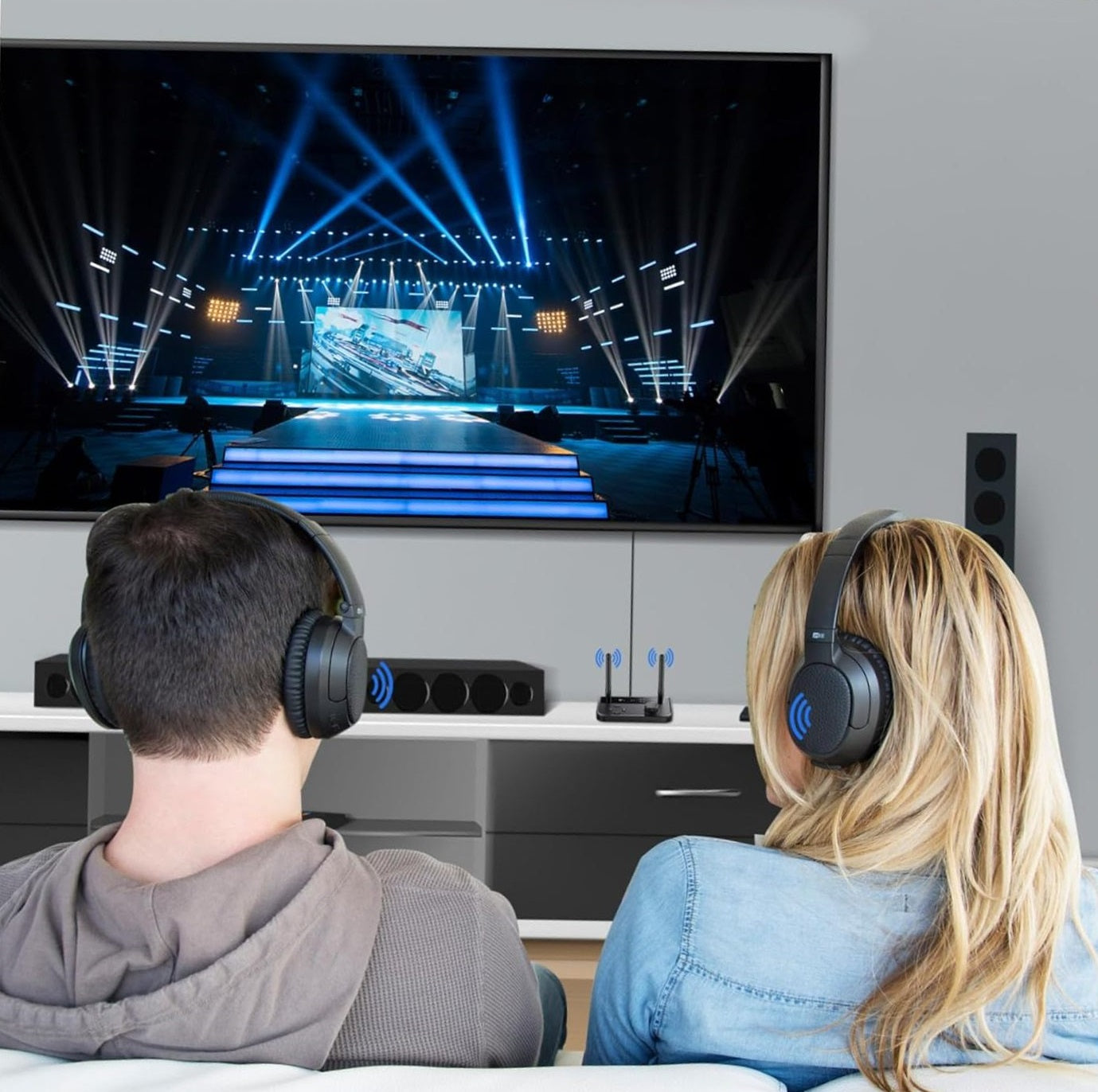 A man and woman wearing Bluetooth headphones, watching a TV connected to the Bluetooth audio transmitter, demonstrating shared private listening.