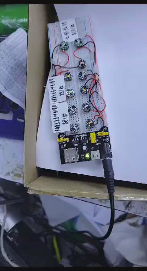 Close-up of a circuit board with LED lights and wiring in a cardboard box, showcasing DIY electronics setup.