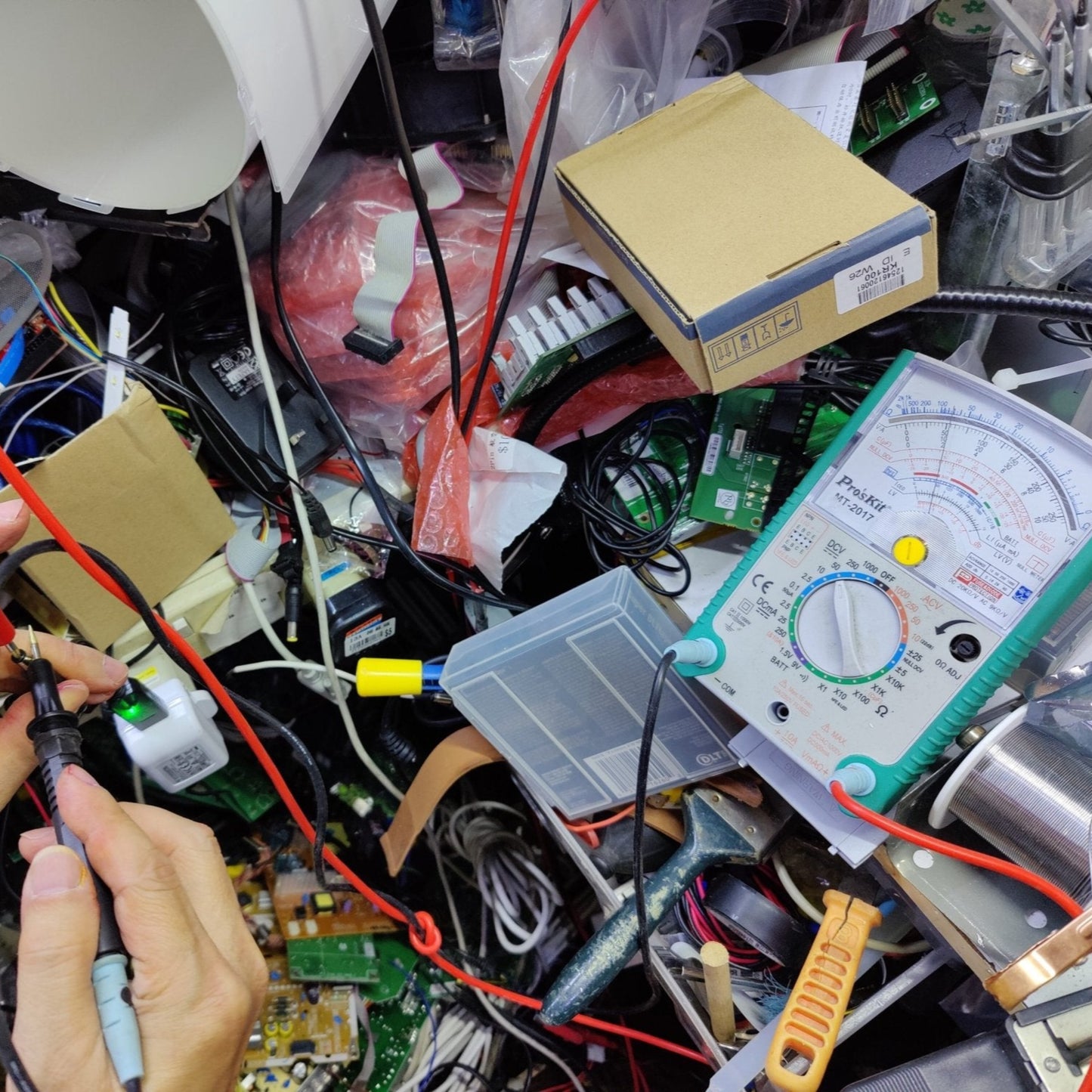 Hands holding multimeter probes amid a cluttered workspace with various electronics and tools.