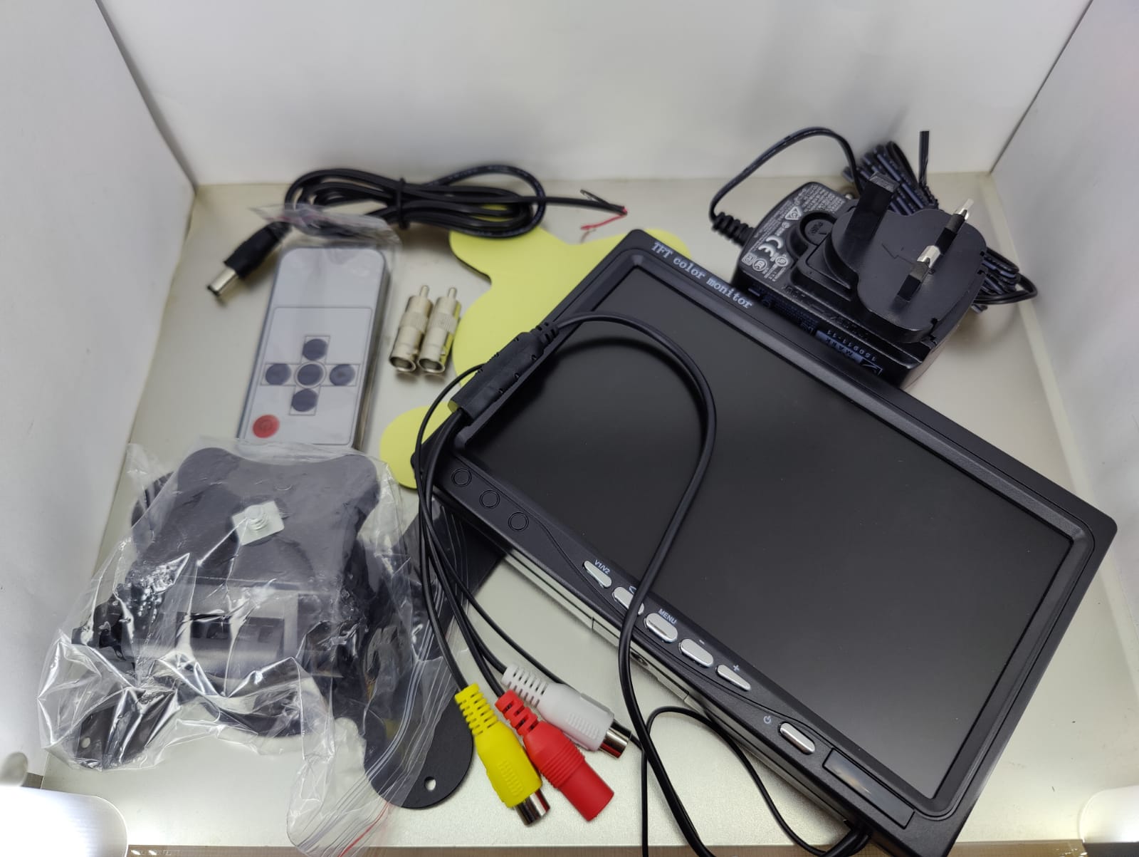 An unboxed 7-inch CCTV monitor is laid out with its accessories, including the mounting bracket, remote control, cables, and power adapter, illustrating the complete package for setting up a security surveillance display.