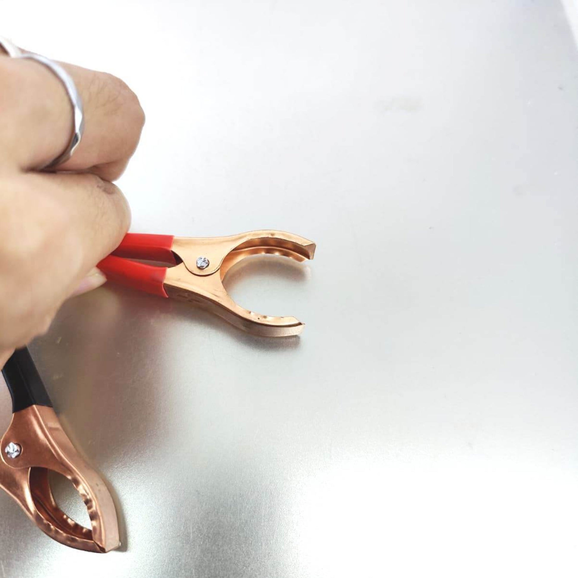 Close-up of a hand holding a red alligator clip, used for electrical connections, on a shiny metallic surface.