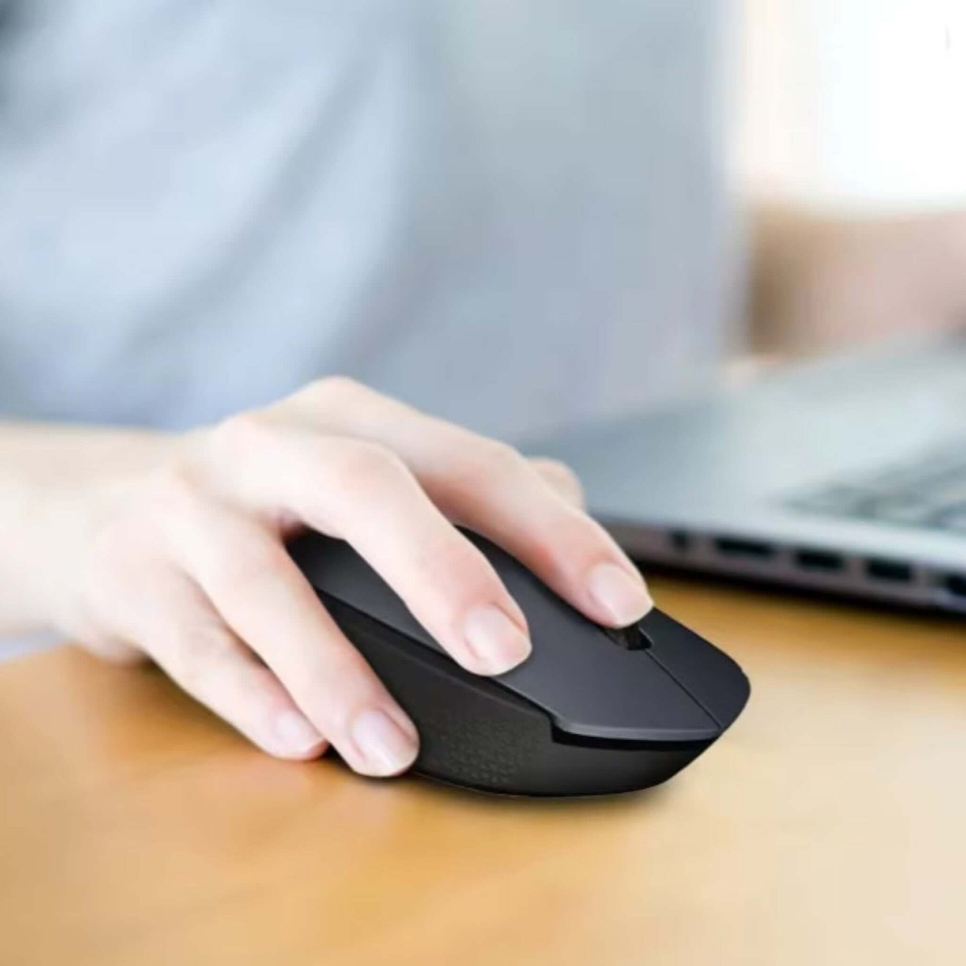 Hand using a dark grey Logitech M170 wireless mouse on a wooden desk with a laptop in the background.