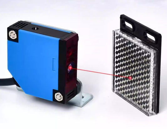 A reflective photoelectric sensor emitting a red laser beam towards a rectangular reflector, demonstrating object detection.