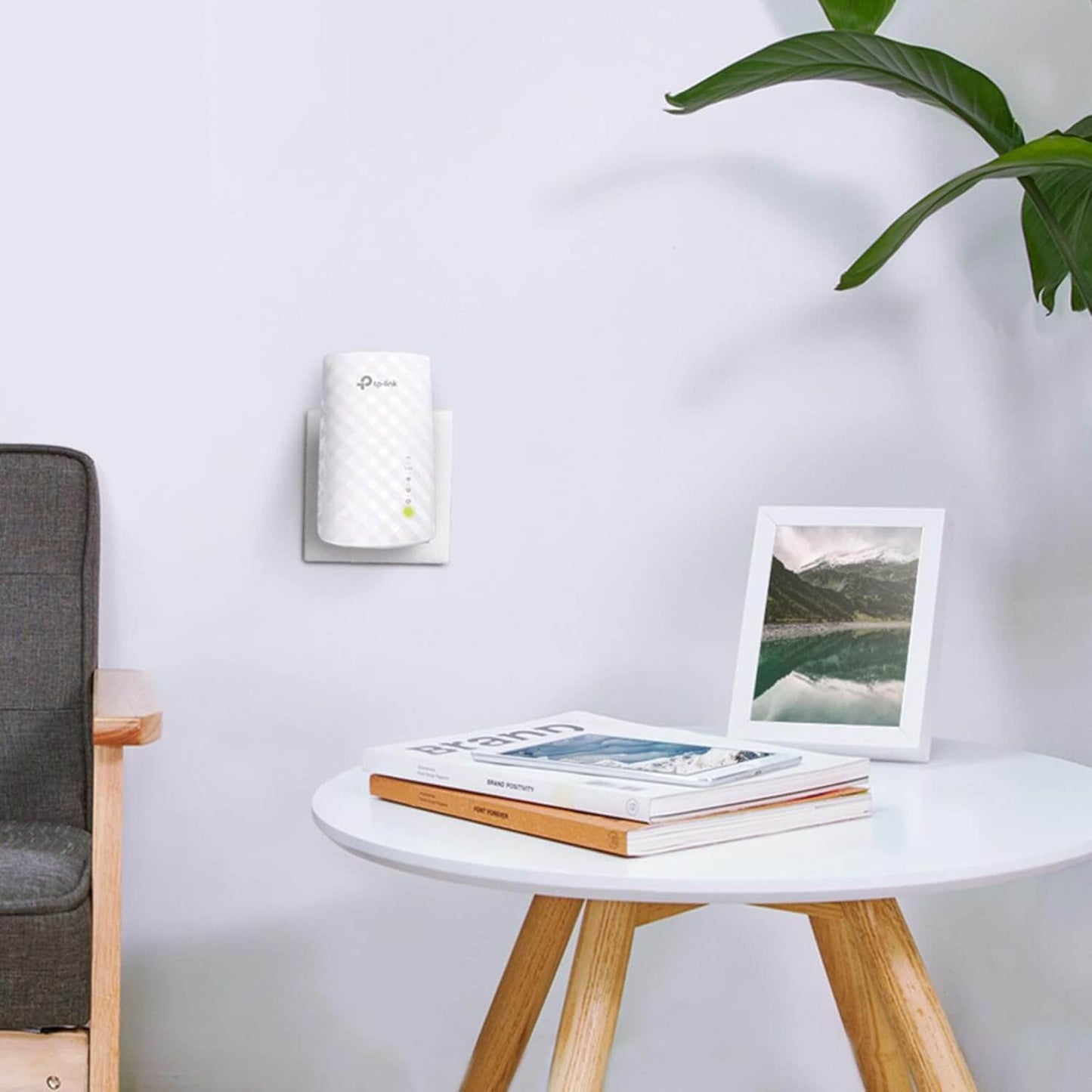 TP-Link RE200 AC750 Dual-Band Wi-Fi Range Extender mounted on wall next to a modern table with magazines and a photo frame.