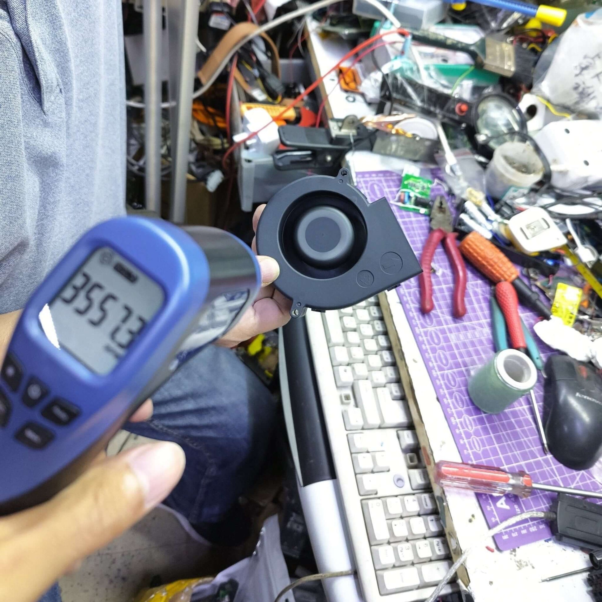 Person measuring temperature with a handheld device in a cluttered workspace with electronic components.