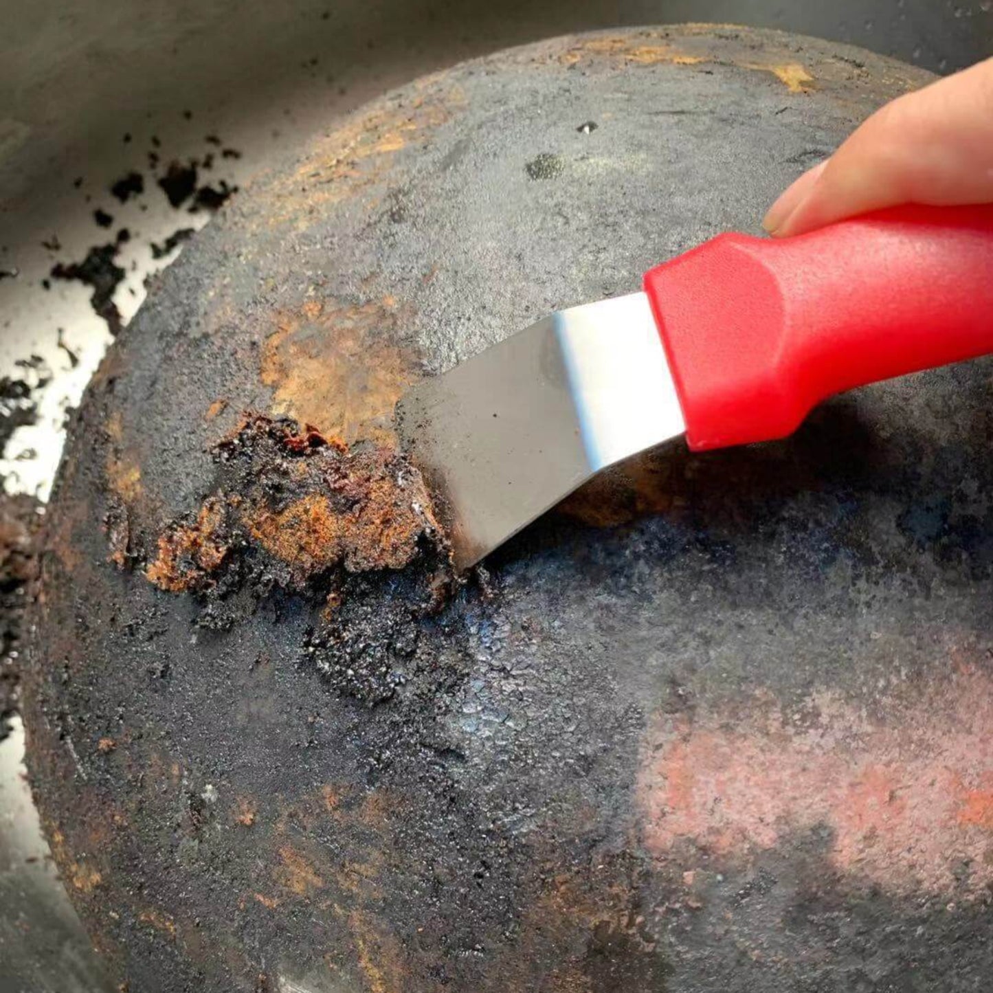 Stainless steel hand scraper with red handle removing burnt residue from a pot in a sink.